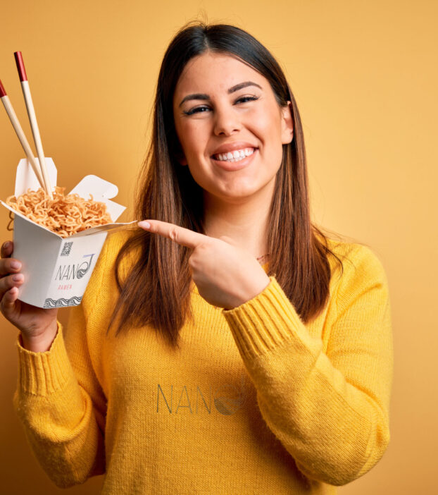 Young beautiful woman eating asian ramen noodles using chopsticks over yellow background very happy pointing with hand and finger